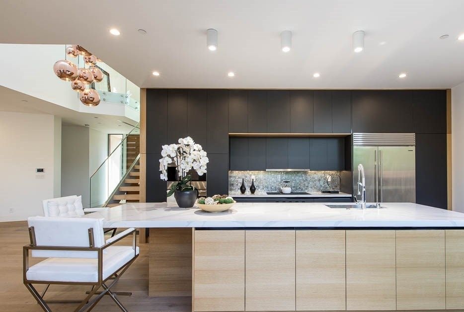 Modern kitchen with long white waterfall island, light wood cabinetry, matte black wall cabinets, and glass stair railing.
